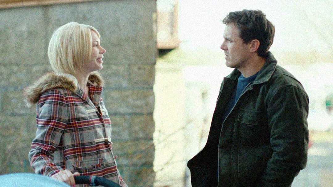 A woman in a plaid coat and a man in a dark jacket stand outside, facing each other and talking, with a stone wall and soft daylight in the background.
