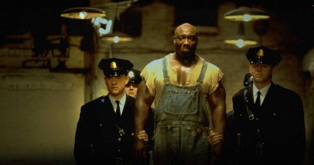 A large man in overalls is escorted by two uniformed police officers down a dimly lit corridor, with hanging lamps above and a somber, tense atmosphere.