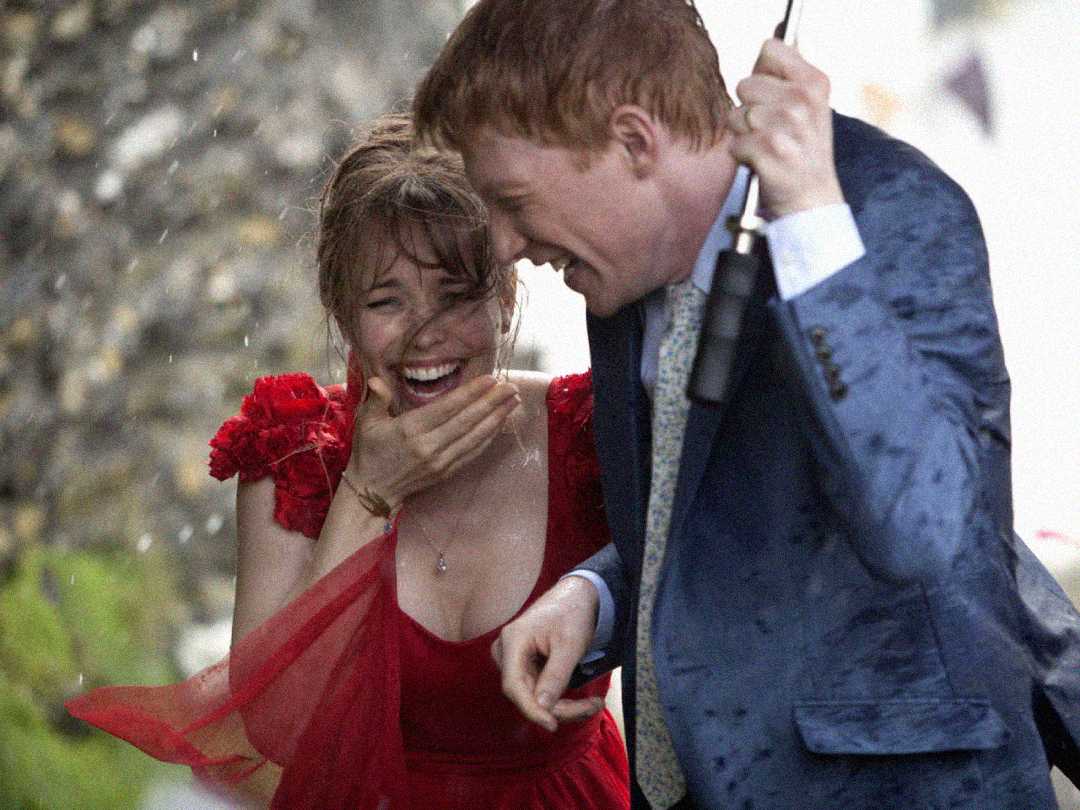A woman in a red dress and a man in a suit laugh together under an umbrella in the rain, looking joyful and soaked, with drops of water visible around them.