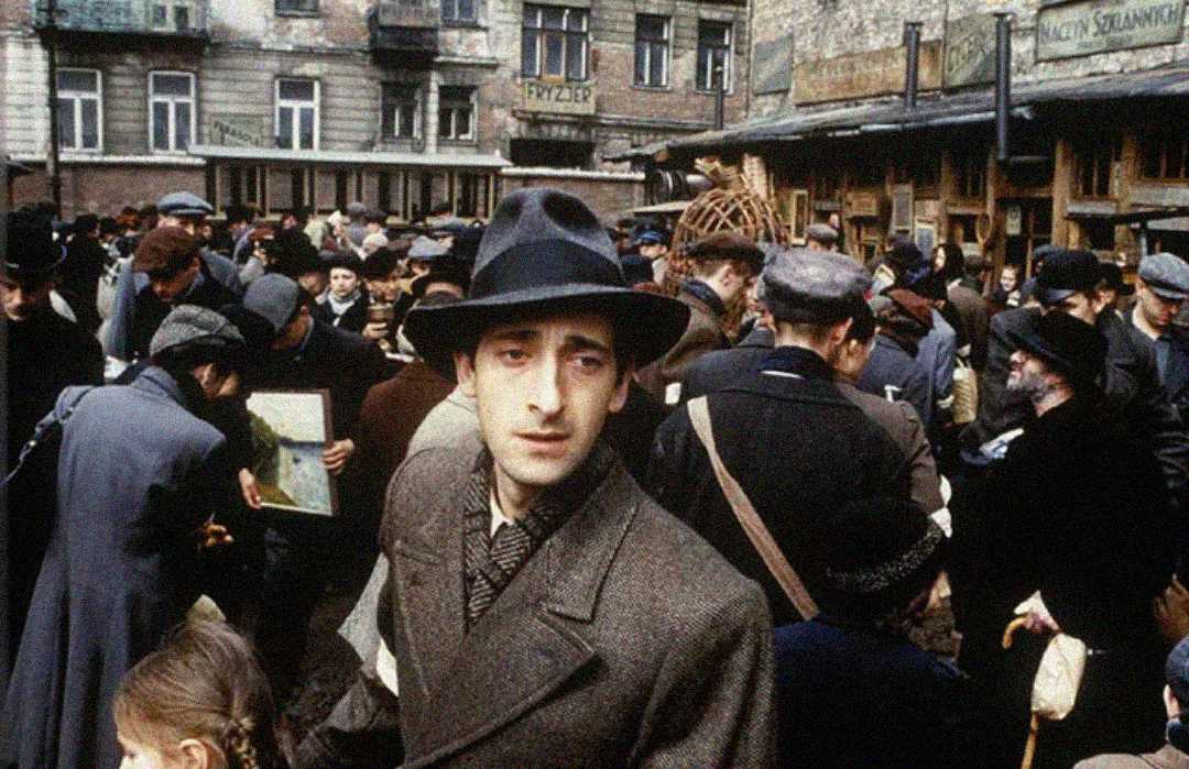 A man in a dark coat and hat stands in the center of a crowded street market, looking directly ahead, as people around him interact, shop, and move through the busy scene. Old buildings appear in the background.