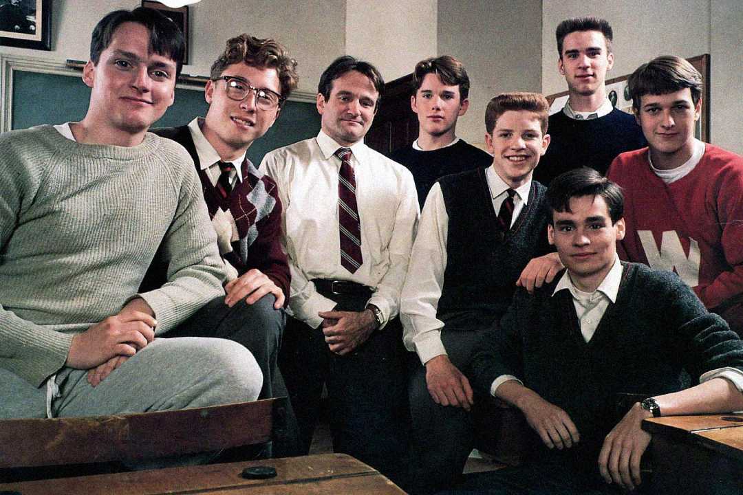 A group of young men and an adult man, wearing sweaters, shirts, and ties, pose together in a classroom setting, smiling at the camera. Desks and neutral walls are visible in the background.