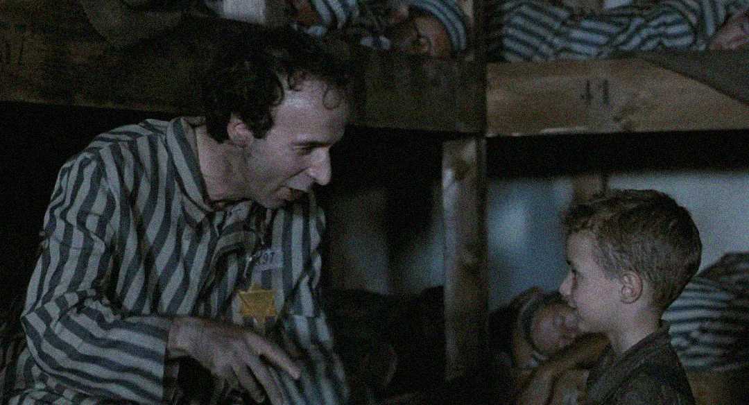 A man and a young boy, both wearing striped concentration camp uniforms with yellow stars, sit together in a dimly lit bunkhouse. The man smiles and gestures while the boy listens, and other prisoners rest in the background.