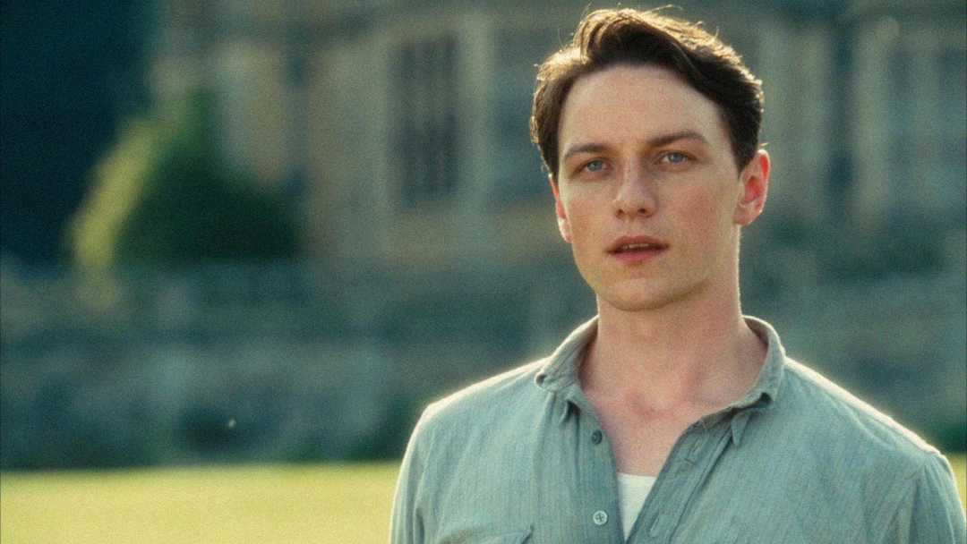 A young man with short brown hair, wearing a light-colored button-up shirt, stands outside in soft sunlight with a blurred building in the background. He looks slightly off-camera with a thoughtful expression.