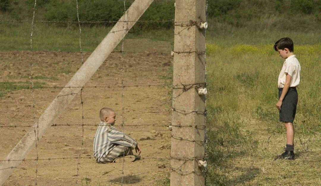 A boy in striped clothing sits behind a barbed wire fence while another boy in shorts and a collared shirt stands on the other side, facing him, with grass and dirt visible in the background.