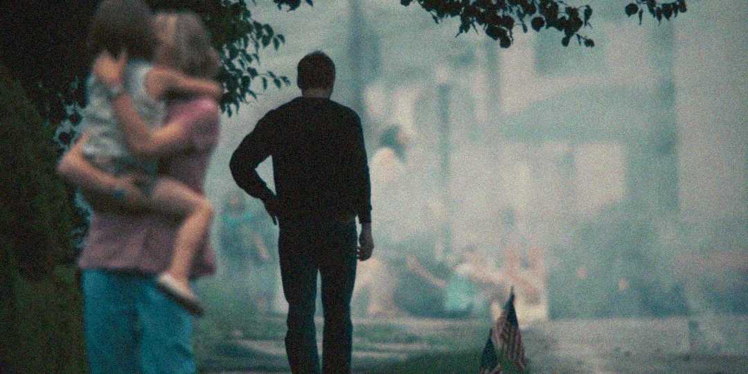 A person carrying a child stands in the foreground, out of focus, while another person walks away down a misty, tree-lined street in the background. The scene appears atmospheric and moody.