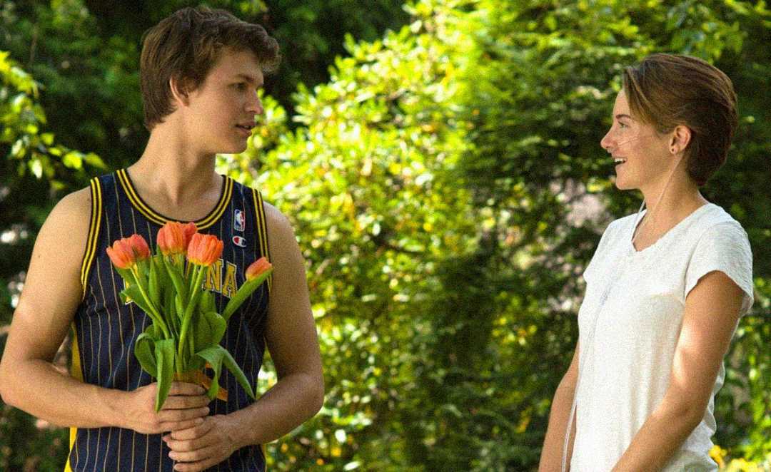 A young man in a basketball jersey holds orange tulips, looking at a smiling young woman in a white shirt. They stand outside with green trees in the background.