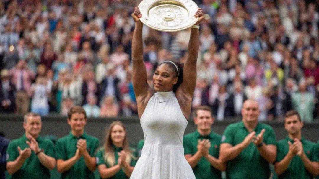 A female tennis player in a white dress holds a large trophy plate above her head on a tennis court, with a cheering crowd and applauding officials in green uniforms behind her.