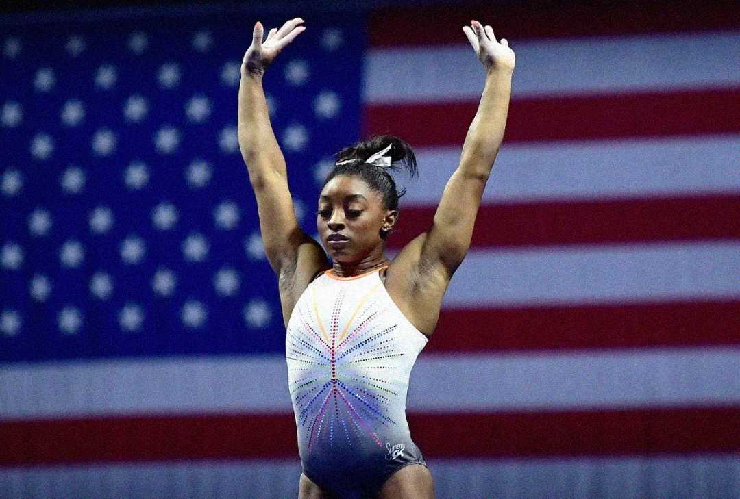 A female gymnast in a leotard stands with both arms raised in front of a large American flag, appearing focused and poised, likely during a competition.