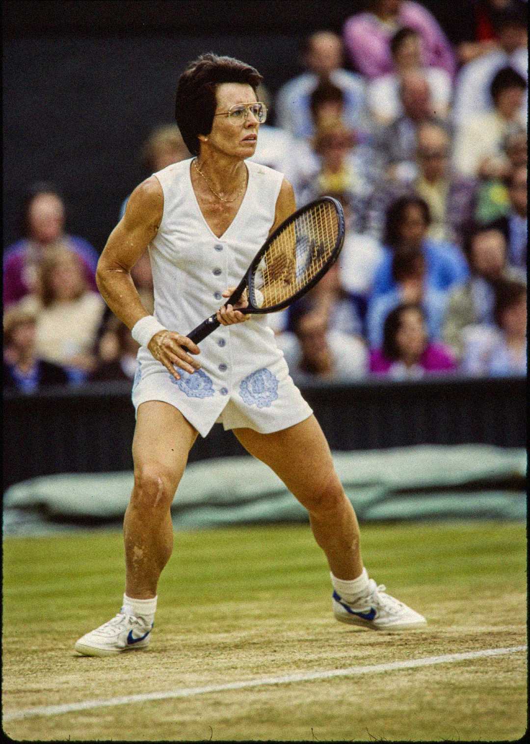 A female tennis player in a white sleeveless outfit is ready in an athletic stance on a grass court, holding a racket with both hands. Spectators are visible in the background.