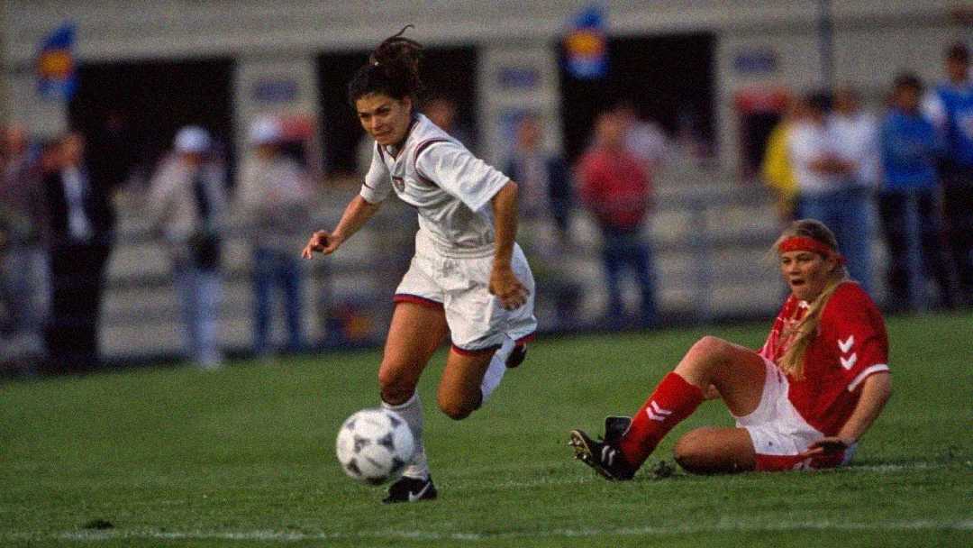 A soccer player in a white uniform runs towards the ball while a player in a red uniform sits on the grass behind her, appearing to have fallen during the match. A crowd is blurred in the background.