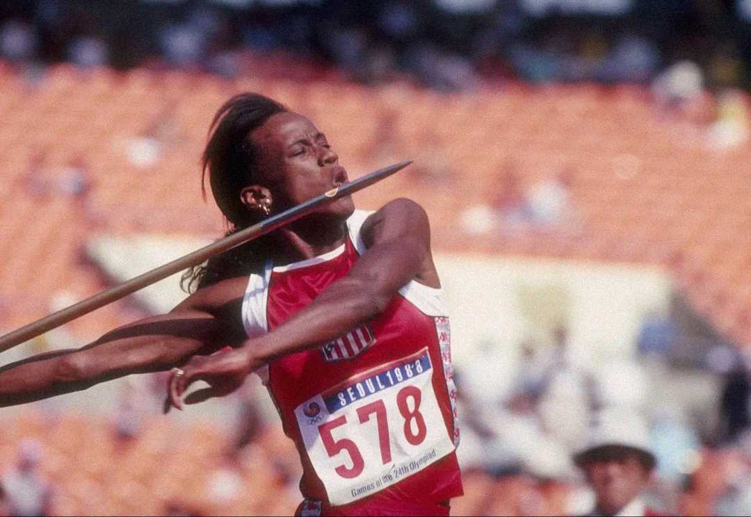 A female athlete wearing a red and white uniform with the number 578 throws a javelin in a stadium during a track and field competition.