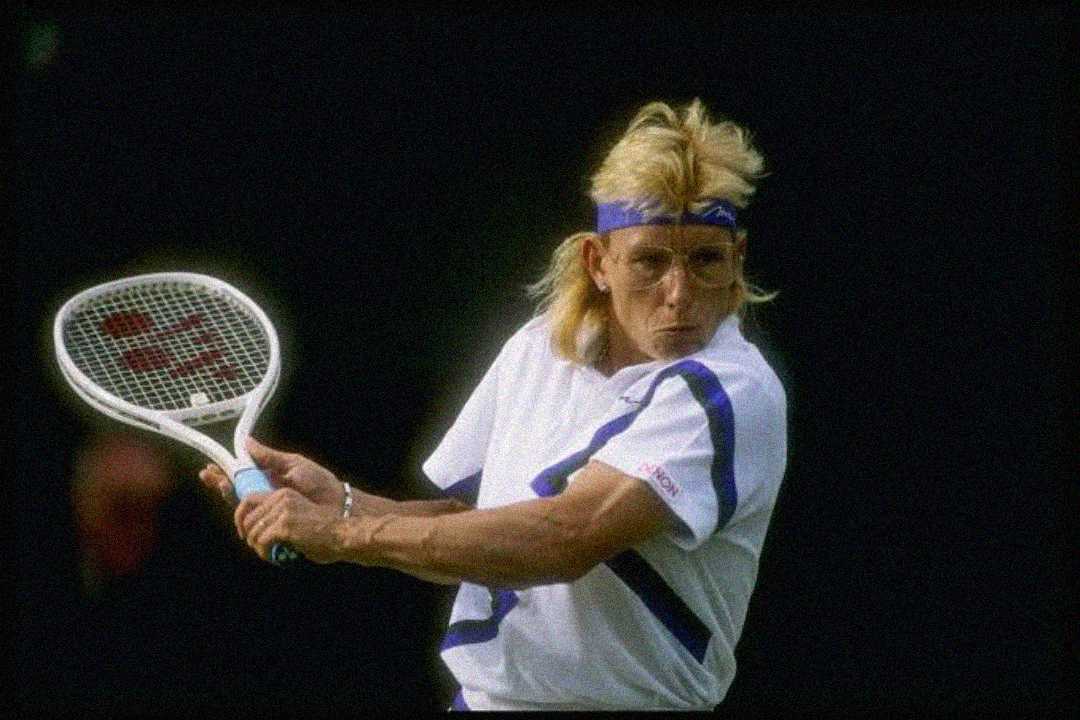 A tennis player with blond hair, wearing a white outfit with purple accents and a purple headband, prepares to hit a backhand shot during a match.