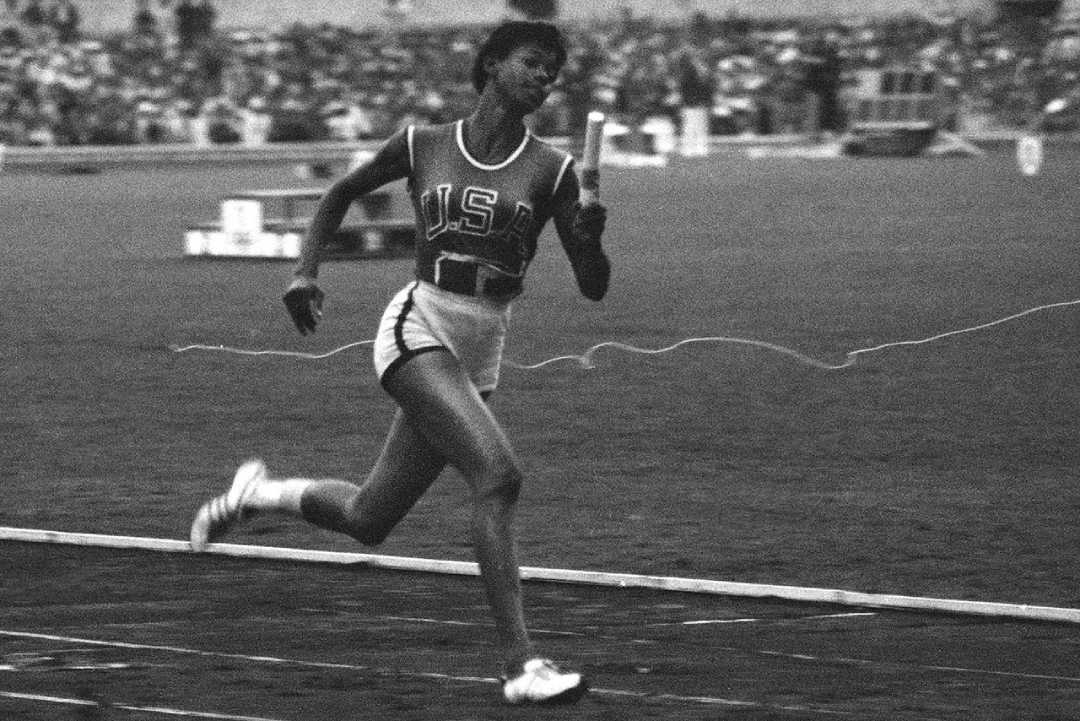 A Black female athlete wearing a "USA" uniform runs on a track with a baton, likely in a relay race, with a blurred stadium crowd in the background. The image is in black and white.