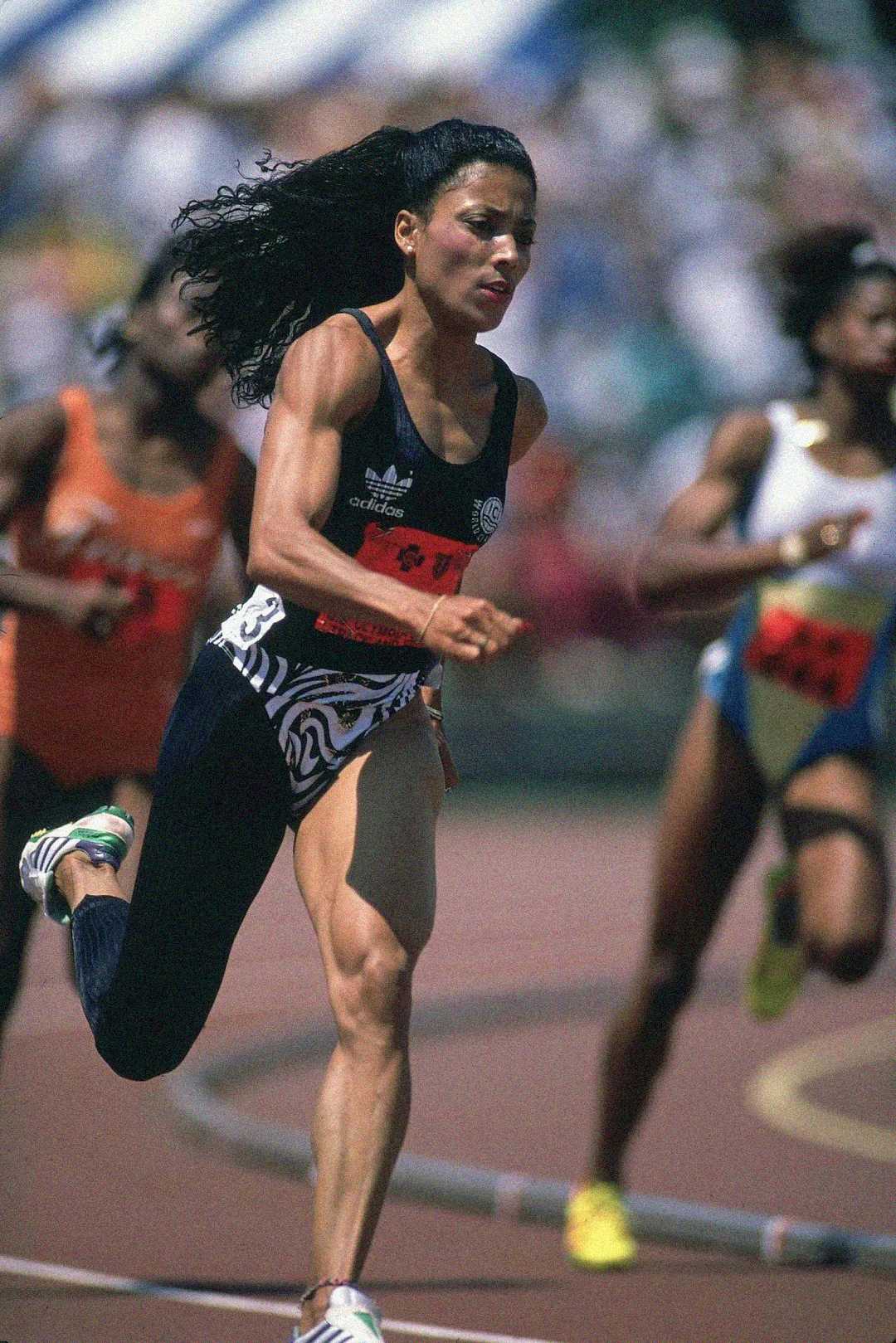 A female sprinter in a black and white uniform runs on a track during a race, with other competitors visible behind her. She appears focused and determined, with her hair flowing back as she sprints.