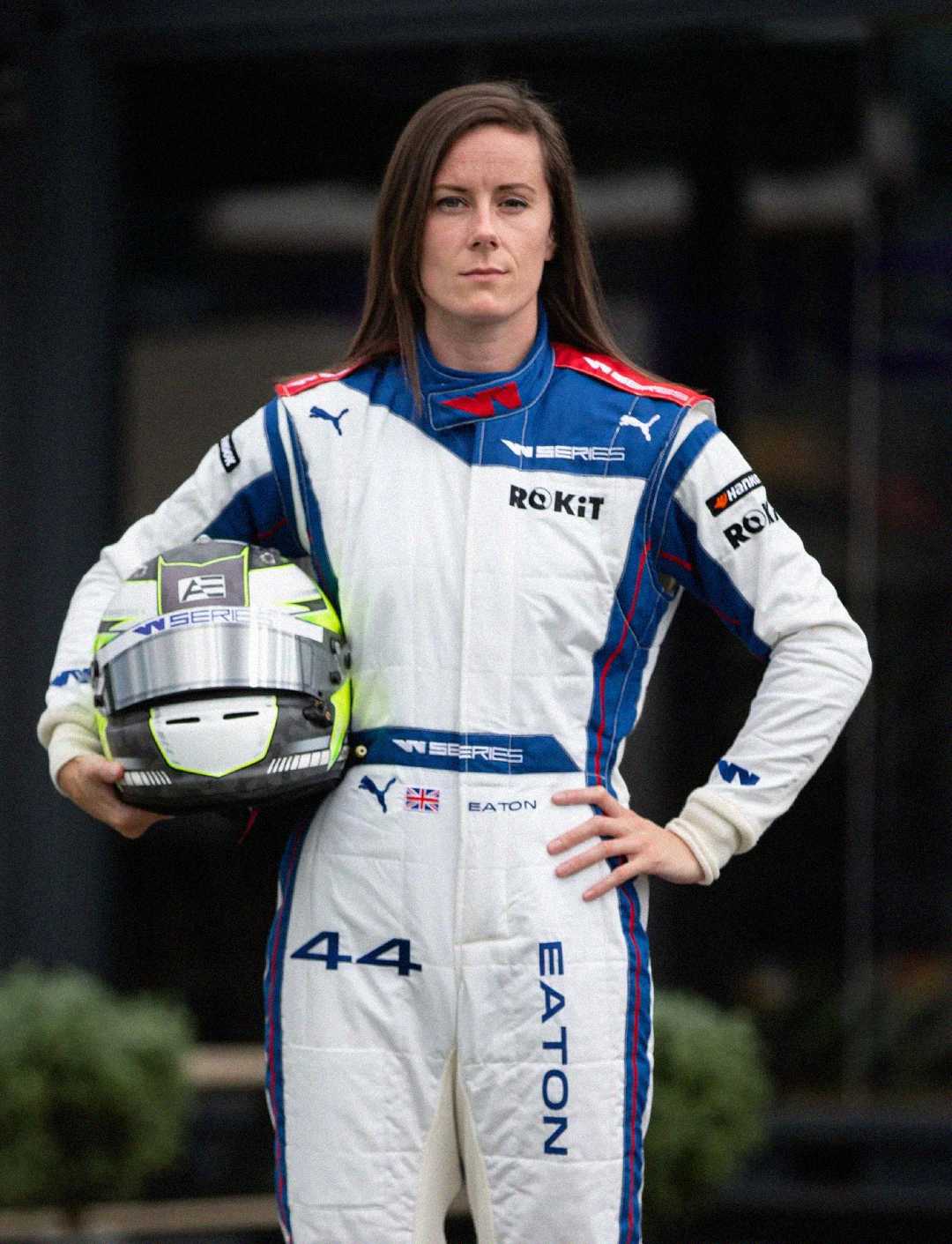 A race car driver in a white and blue racing suit stands confidently, holding a helmet under one arm. The suit features sponsor logos, the number 44, and the name "EATON." The background is blurred.