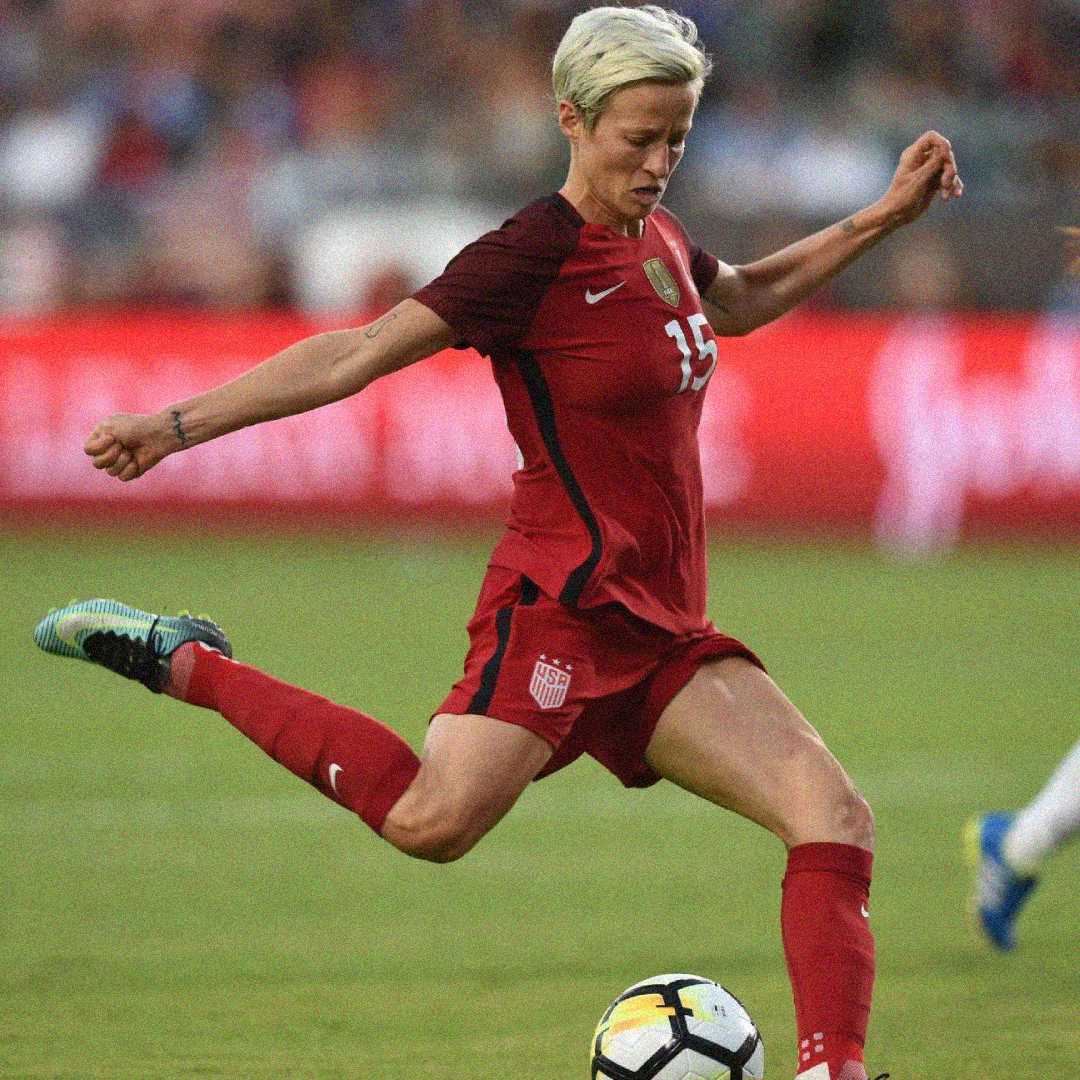 A soccer player in a red USA team uniform with the number 15 is about to kick a soccer ball during a game on a grassy field. The background shows a blurred crowd and a red advertisement banner.