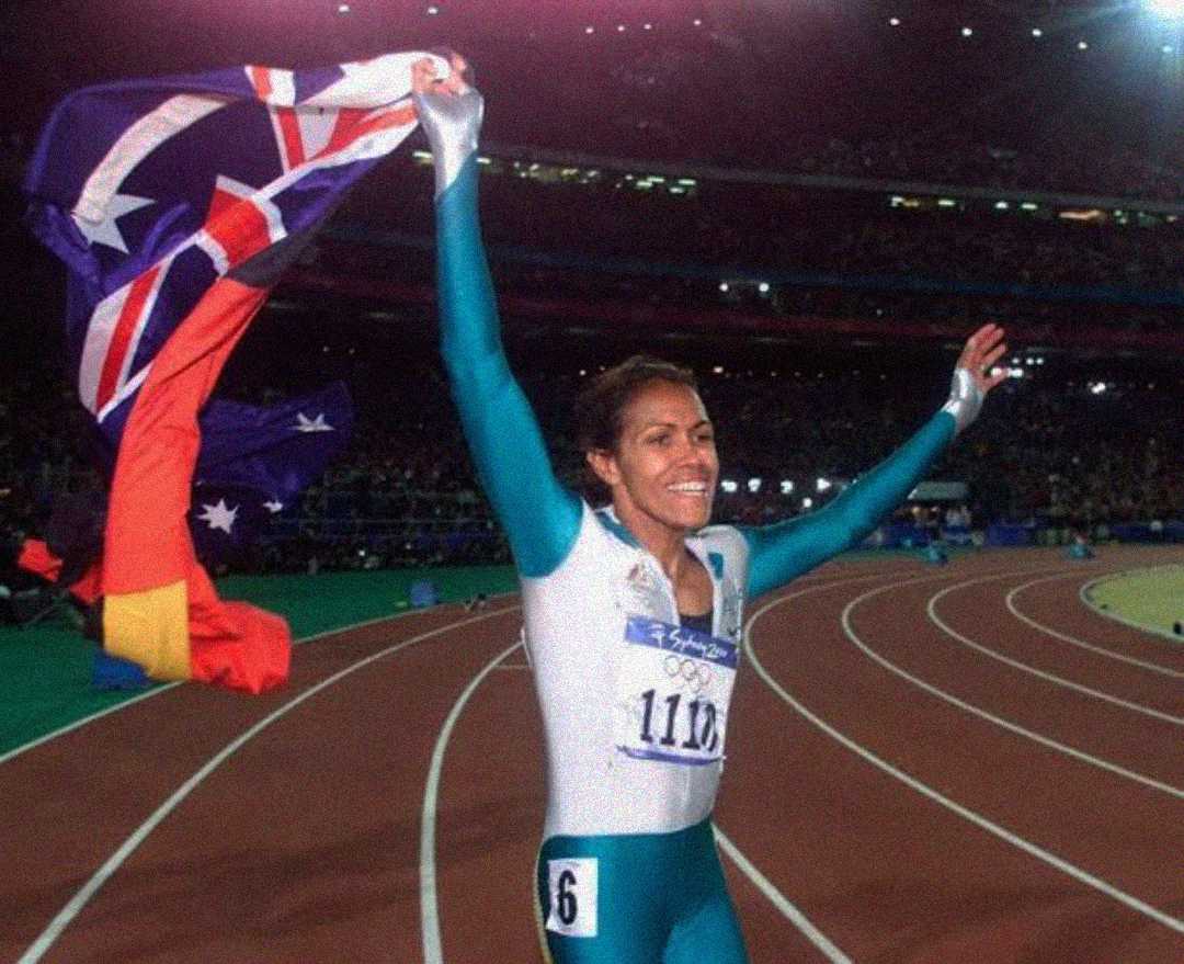 A female athlete in a green and white uniform celebrates on a track, smiling and holding up an Australian flag during a nighttime stadium event.