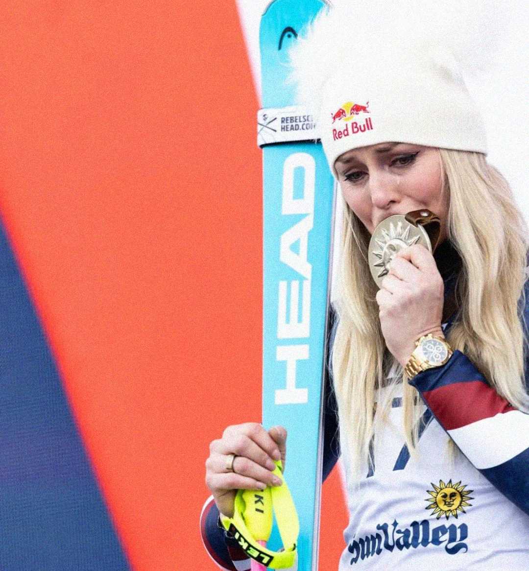 A female skier wearing a white beanie and ski gear holds a turquoise HEAD ski and bites a silver medal, appearing emotional during an award ceremony.