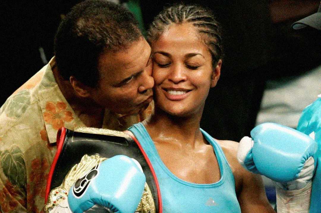A man kisses a smiling female boxer on the cheek. She is wearing blue boxing gloves and holding a championship belt, appearing happy and victorious in the boxing ring.