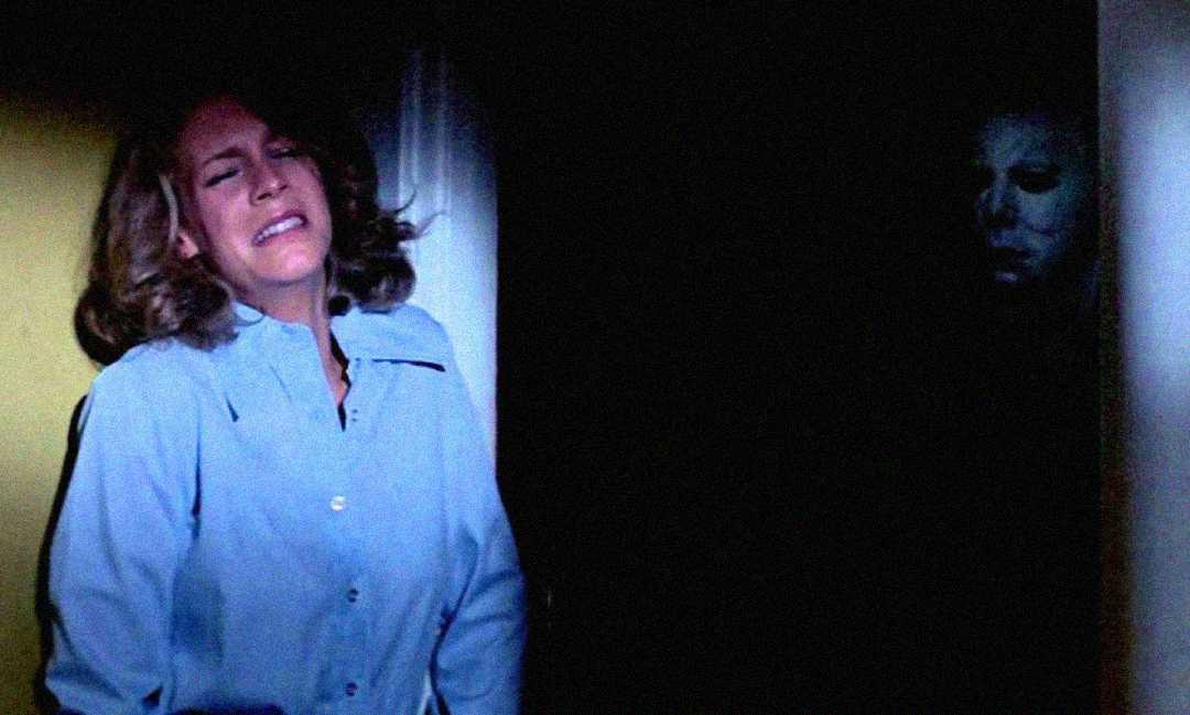 A frightened woman in a blue shirt stands against a lit wall, while a figure in a white mask lurks ominously in the dark background.