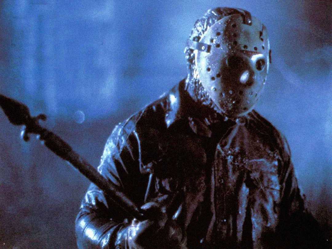A person wearing a worn hockey mask and dark, tattered clothing stands in dim blue lighting, holding a large spear-like weapon. The scene appears eerie and suspenseful.
