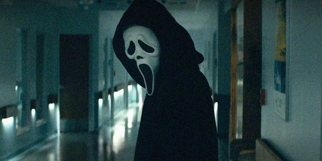 A person wearing a black hooded cloak and a white Ghostface mask stands in a dimly lit hallway, creating a suspenseful and eerie atmosphere.