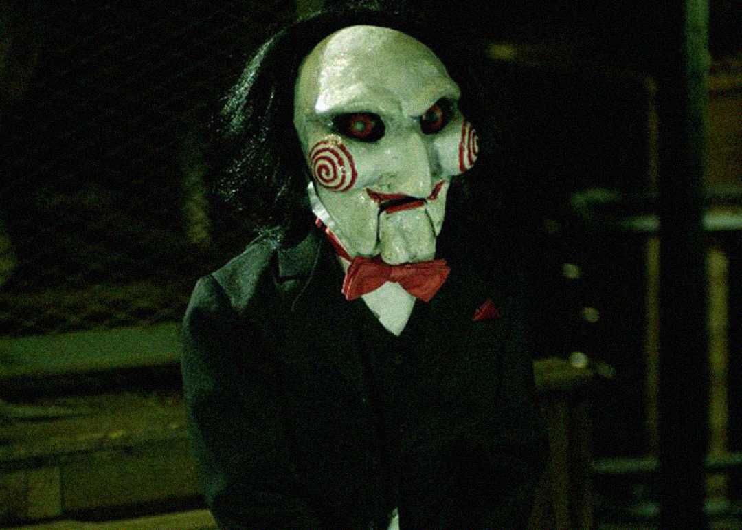 A creepy puppet with white skin, red spiral cheeks, black hair, and a red bow tie, dressed in a black suit, sits in a dimly lit, shadowy room.