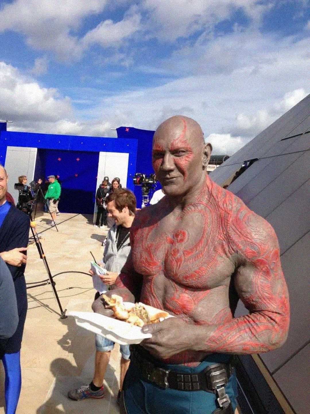 A muscular man in detailed red and gray body makeup, wearing blue pants, holds a plate of food on a film set outdoors. Crew members and equipment are visible in the background under a partly cloudy sky.
