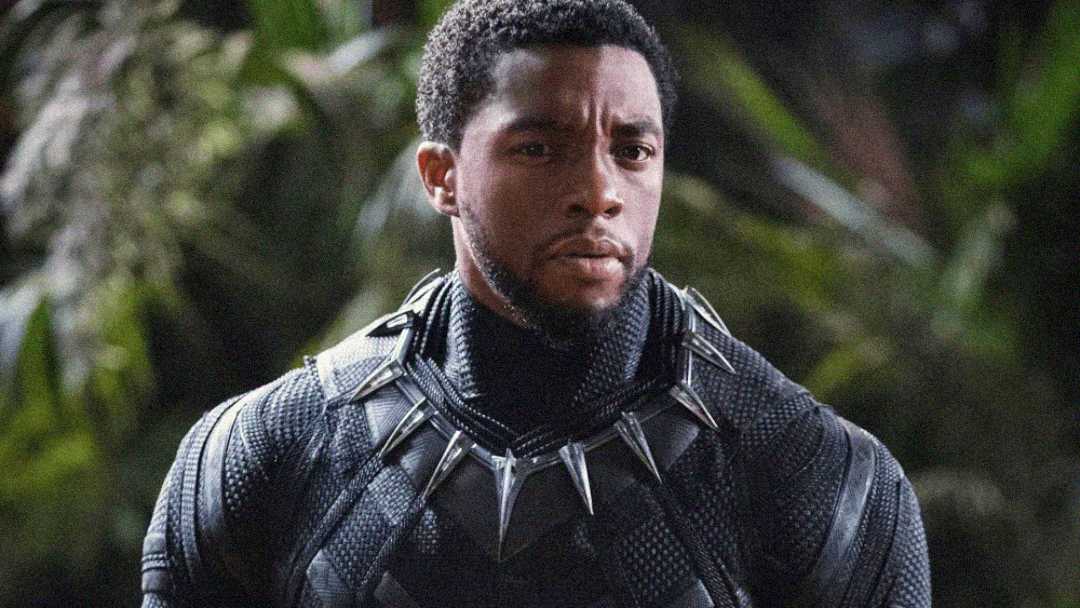 A man wearing a black superhero suit with silver accents and claw necklace stands outdoors, looking serious, with a blurred background of green foliage.