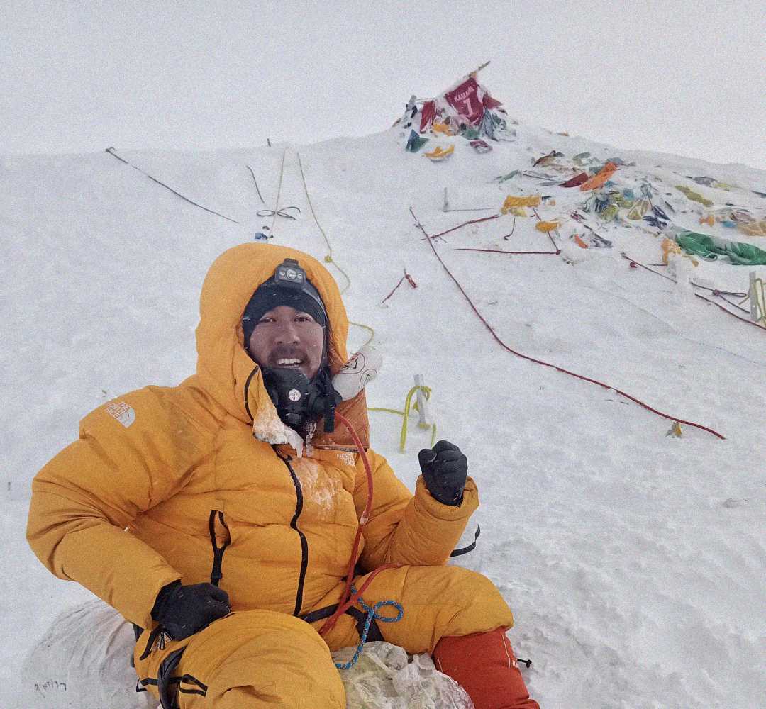 A person in a yellow insulated suit sits on snowy ground, smiling with an oxygen mask, surrounded by climbing ropes and colorful prayer flags on a snowy mountain slope.