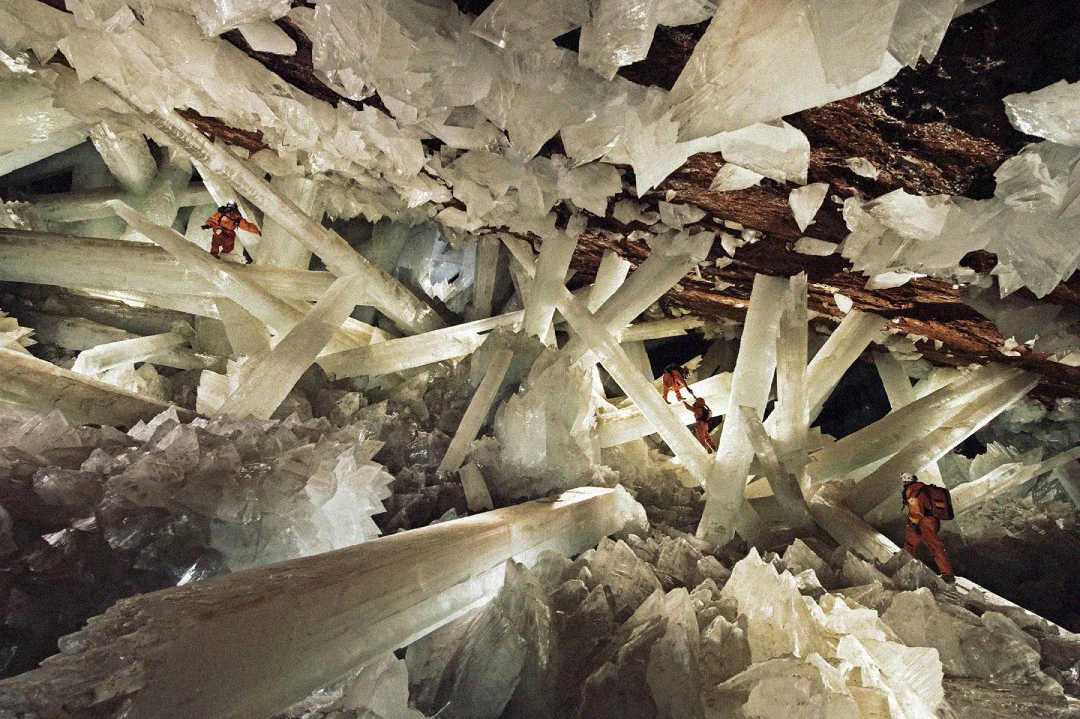 Giant selenite crystals fill an underground cave, with three people in protective suits exploring and climbing among the towering, translucent crystal formations.