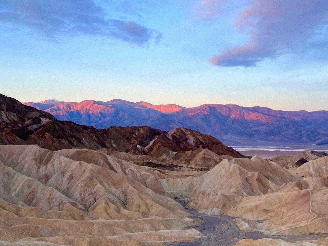 Colorful desert hills and rugged mountains are bathed in the warm light of sunrise or sunset under a partly cloudy sky, with a valley visible in the distance.