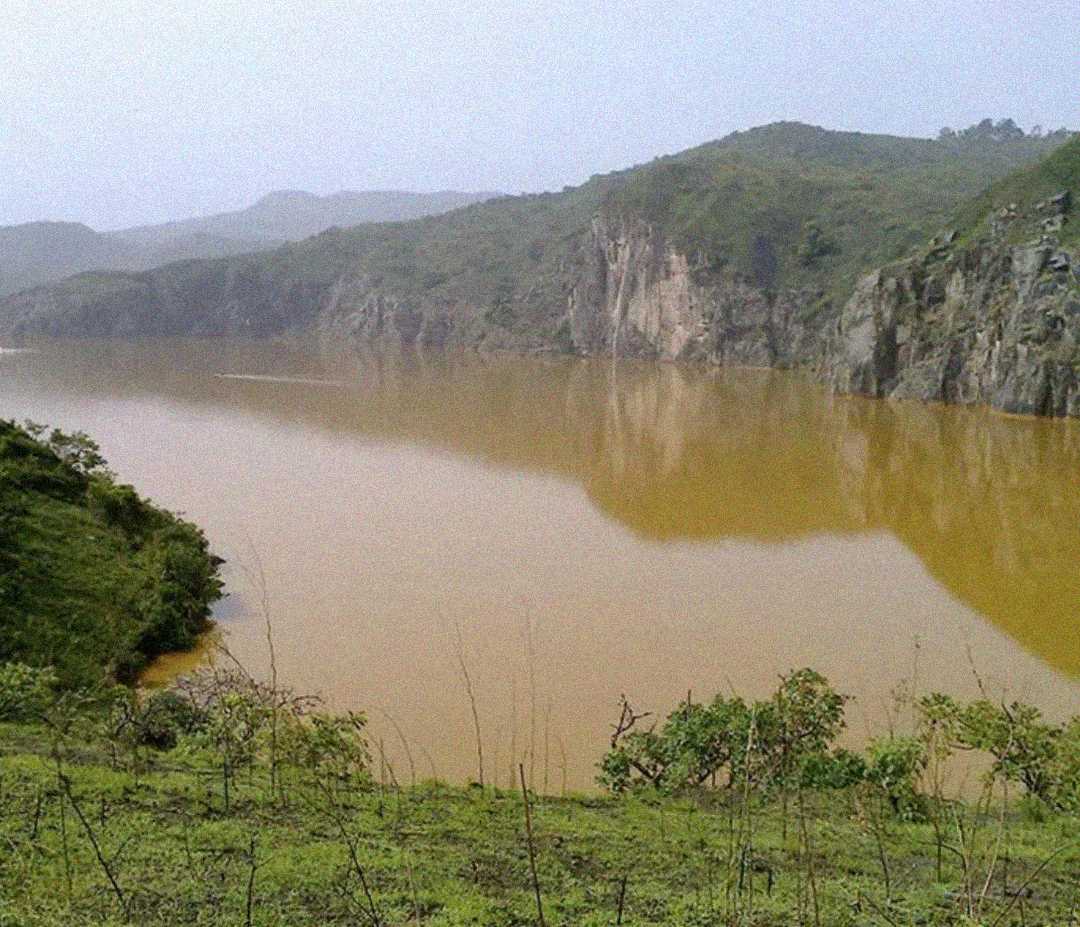 A muddy river winds through green hills and rocky cliffs under a hazy sky, with sparse vegetation in the foreground and mountains in the background.