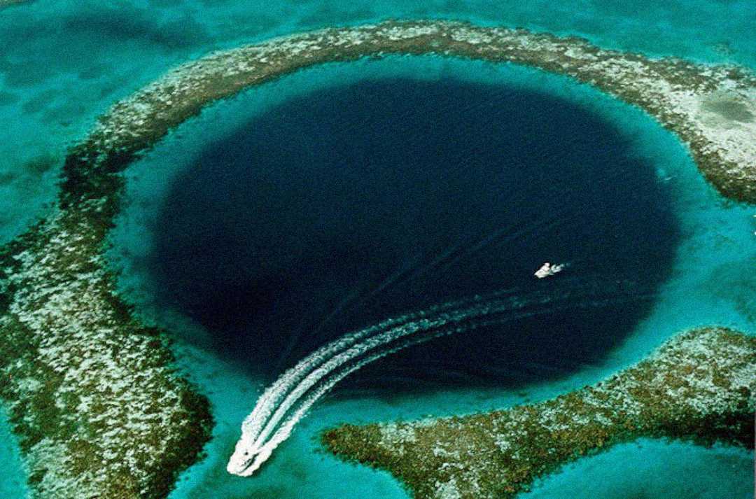 Aerial view of a deep, circular blue hole surrounded by shallow turquoise water and coral, with two boats leaving white trails across the surface.