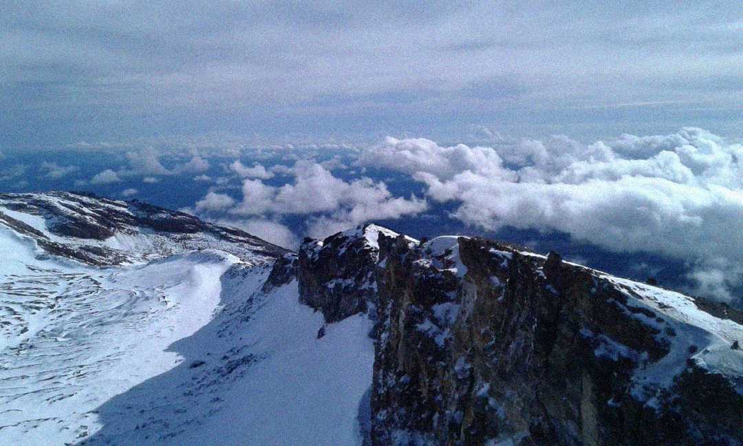 Snow-covered mountain ridge under a cloudy sky, with clouds floating below the peaks and rugged, rocky edges visible along the slope.
