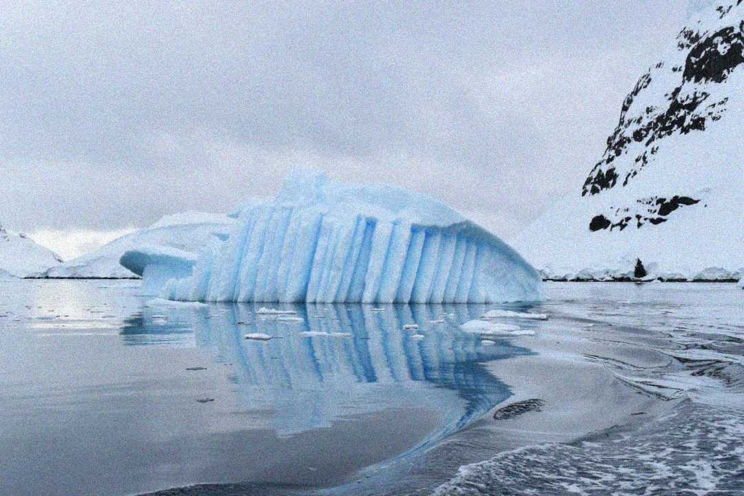 A large iceberg with vertical ridges floats on calm, reflective water, surrounded by snowy mountains under a cloudy sky.