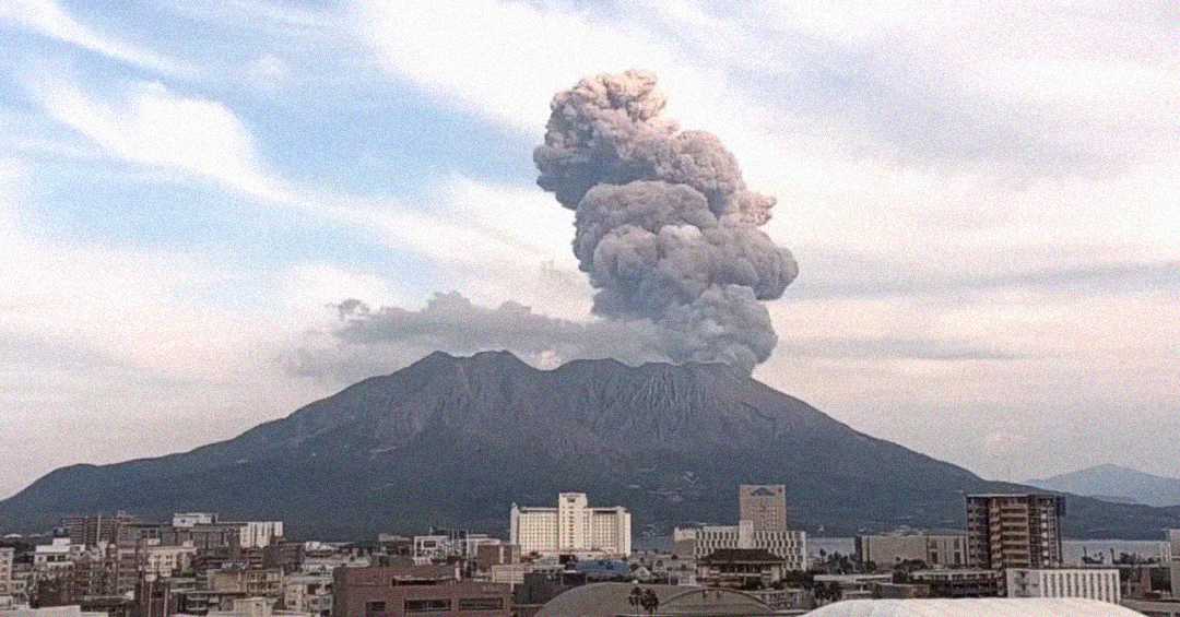 A large volcano emits a thick plume of gray smoke into the sky above a city with various buildings and a mostly cloudy sky in the background.