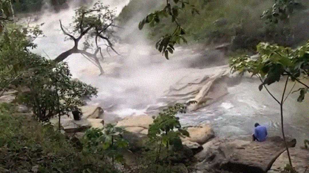 A person sits on a rock beside a steaming river surrounded by lush greenery and trees, with mist rising from the water in a natural setting.