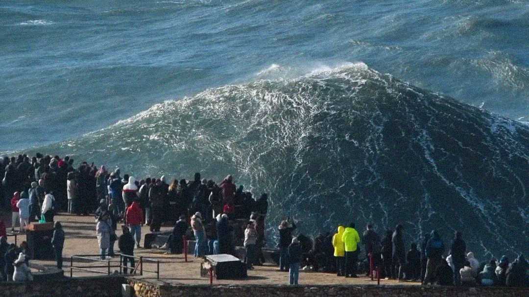 A large crowd stands on a pier or lookout point, watching a massive wave rise dramatically close to them in the ocean. One person in a bright yellow jacket stands out among the onlookers.