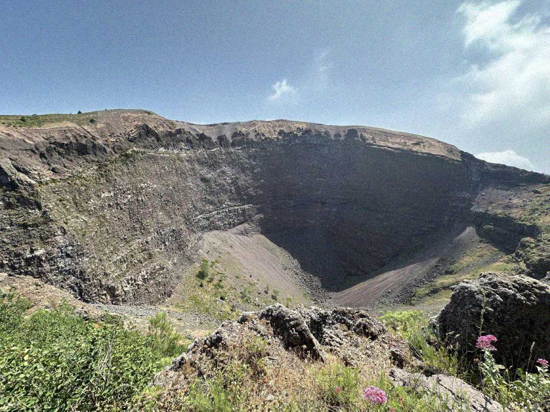 A wide, rocky volcanic crater with steep walls, surrounded by sparse greenery and wildflowers under a blue sky with a few clouds.
