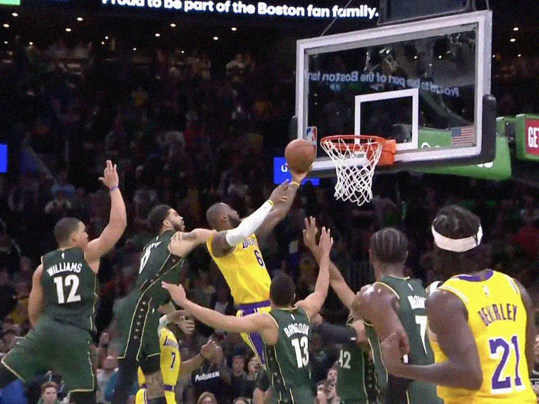 A basketball player in a yellow jersey attempts a layup as several players in green jerseys try to block him near the hoop during an intense game, with the crowd watching in the background.
