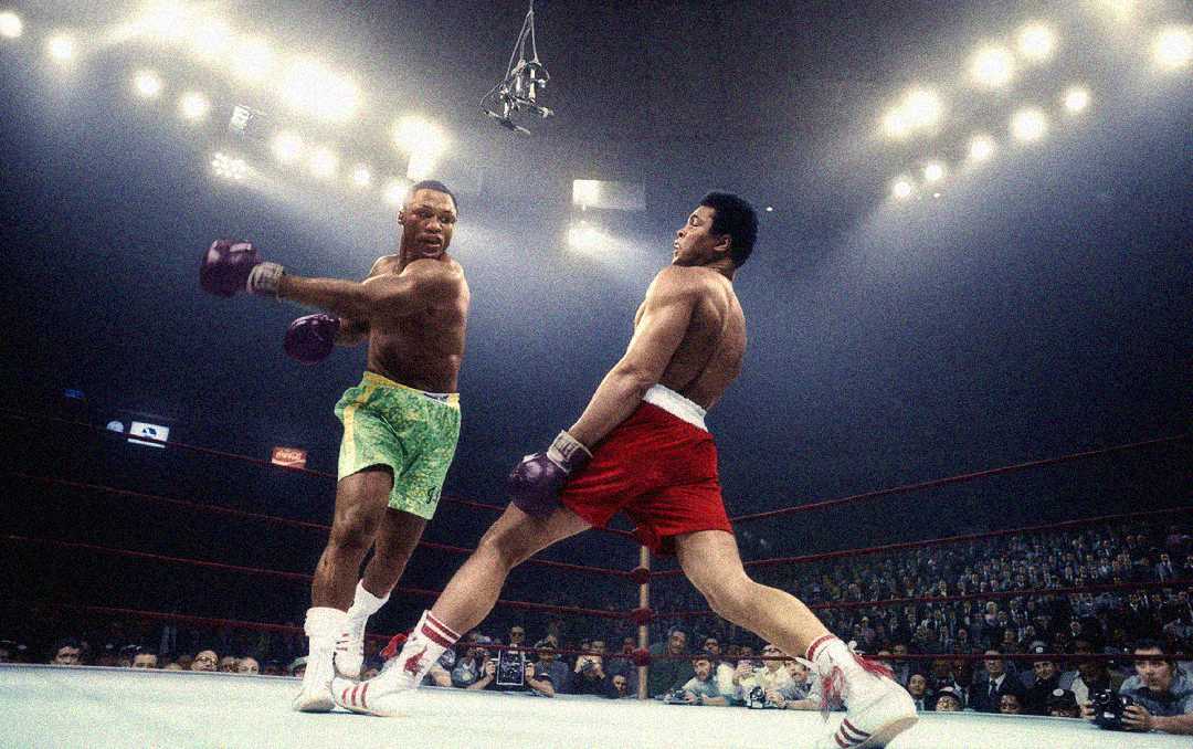 Two male boxers in a brightly lit boxing ring; one in green shorts and purple gloves throws a punch as the other in red shorts leans back to dodge. The audience watches from the stands.