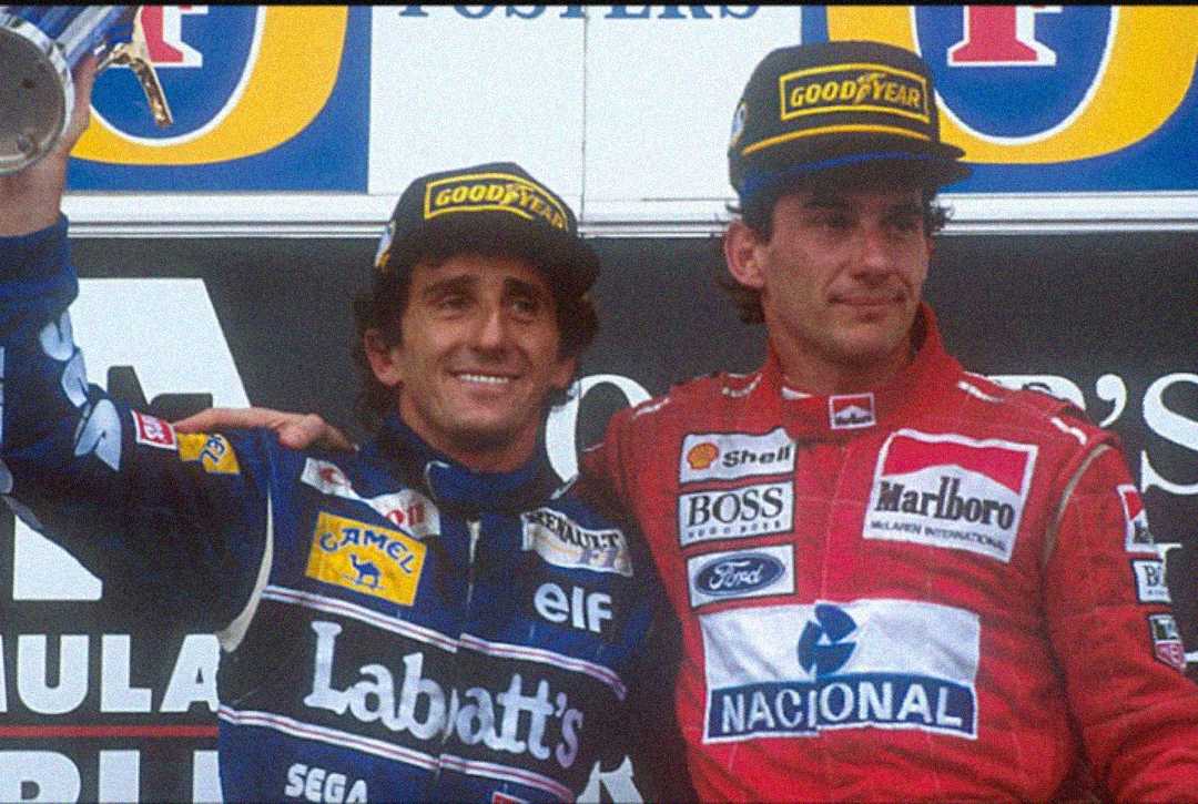 Two Formula 1 drivers wearing racing suits and Goodyear caps stand side by side on a podium, one holding a trophy and smiling, the other with his arm around him, in front of a backdrop with sponsor logos.