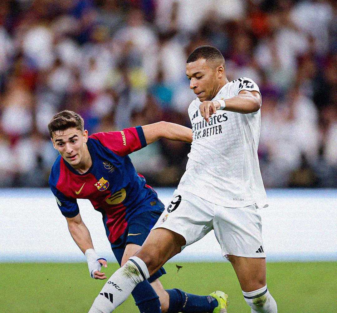 Two soccer players compete for the ball during a match, one in a white kit kicking the ball and the other in a red and blue kit attempting to defend. The background is blurred with spectators.
