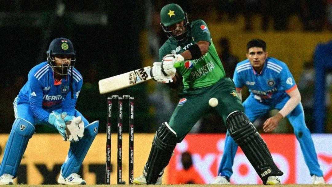A Pakistani cricketer bats while an Indian wicketkeeper and fielder watch closely during a cricket match. The players are in action on the field, wearing their respective team uniforms.