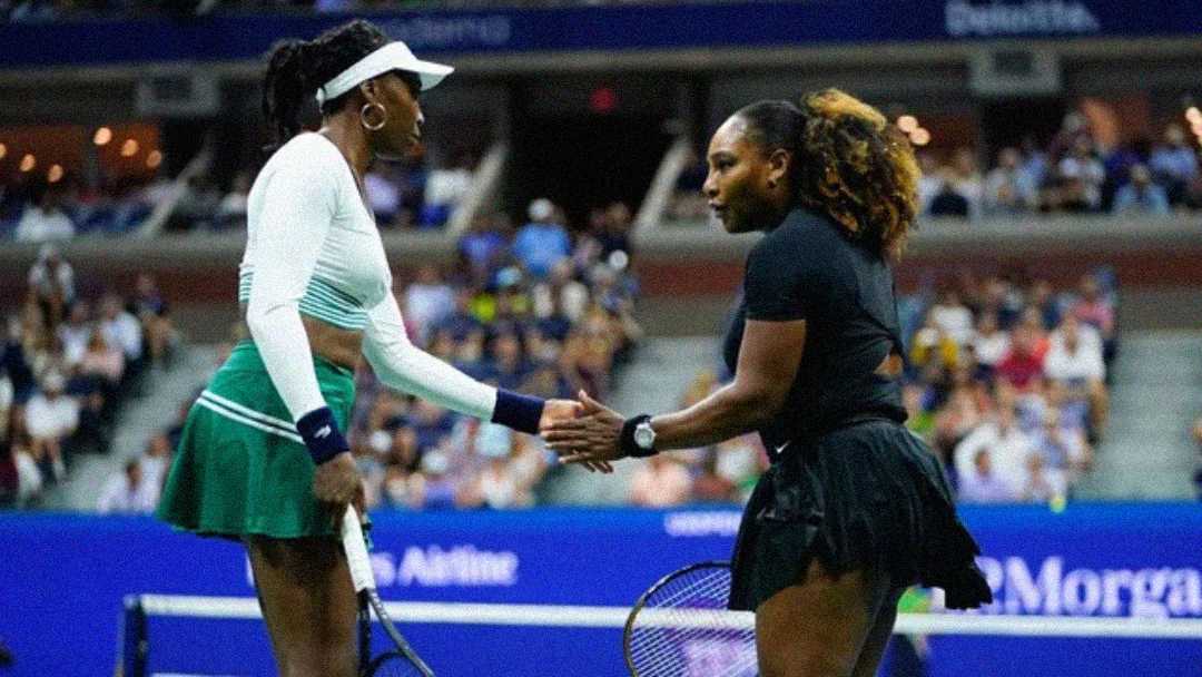 Two female tennis players shake hands across the net after a match on a blue court, with a crowd watching in the background. Both are wearing skirts, visors, and holding tennis rackets.