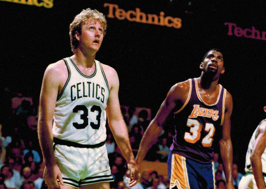 Two basketball players stand on the court during a game. One wears a white Celtics jersey with the number 33, and the other wears a purple Lakers jersey with the number 32. Both look upward, focused.