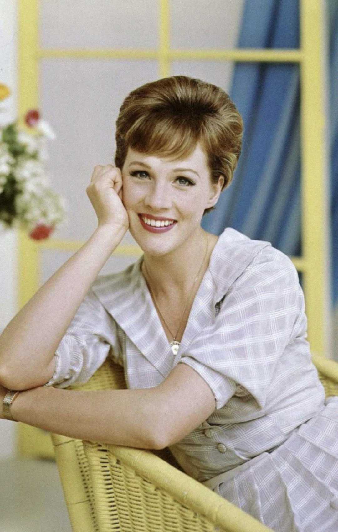 A smiling woman with short brown hair sits on a yellow wicker chair, resting her head on her hand. She wears a light plaid dress and is posed in front of a window with blue curtains and a vase of flowers in the background.