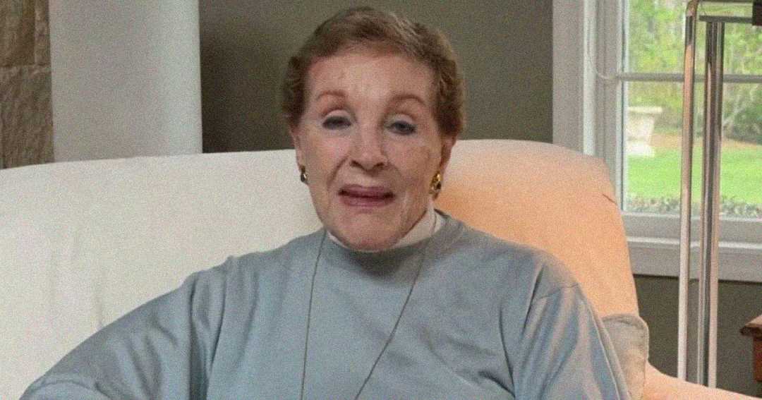 An older woman with short brown hair sits on a light-colored chair indoors, wearing a gray top and necklace. A window with greenery outside is visible in the background.