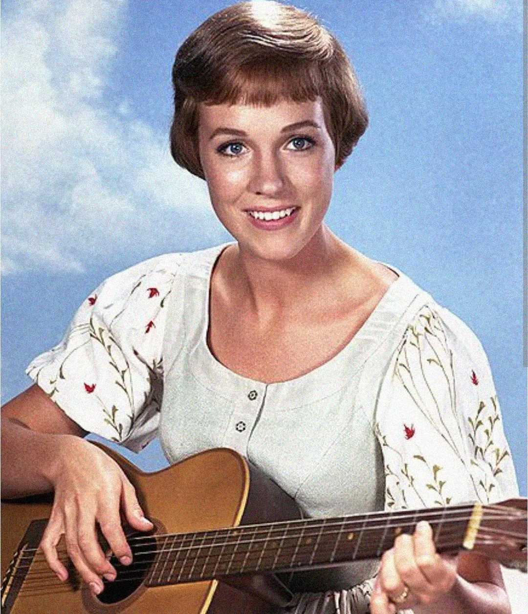 A woman with short brown hair, wearing a light-colored dress with floral embroidery, smiles while playing an acoustic guitar against a blue sky background.